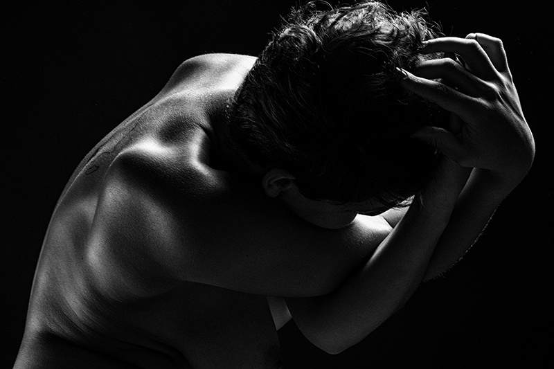 A naked back and head in low light, the arms twisted around each other toward the head.