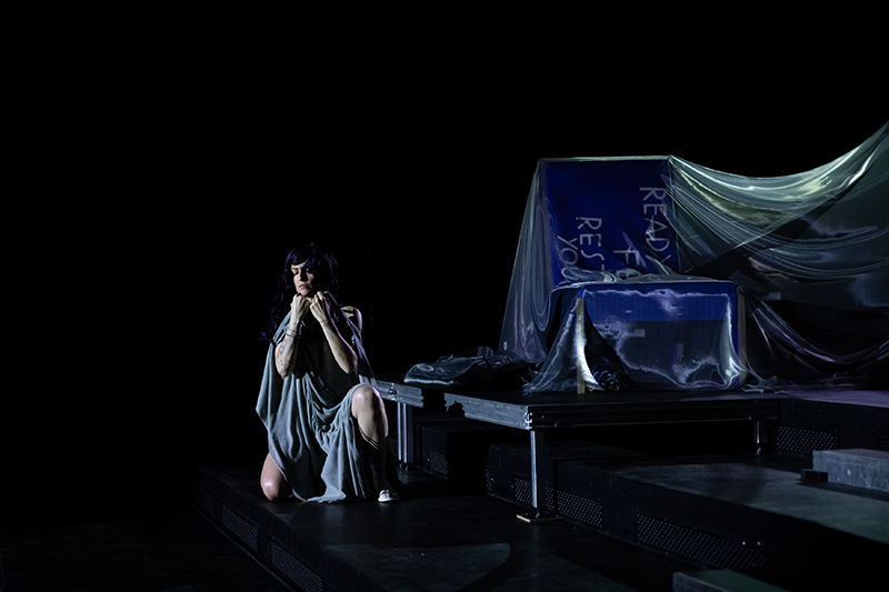 A lone performer in a draped costume kneels on a dimly lit stage beside fabric‑covered set pieces, creating a moody and contemplative atmosphere.