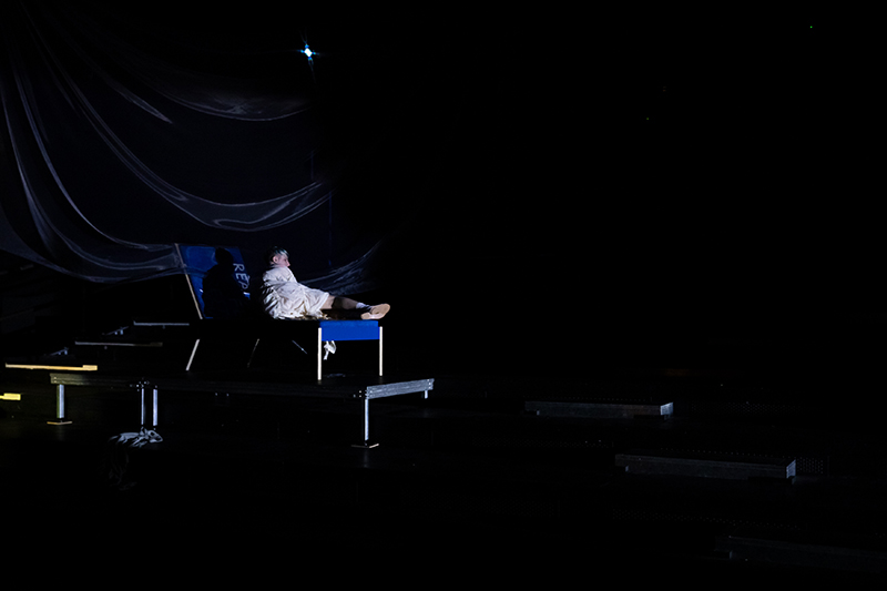 A single performer reclines on a small platform under sparse, moody lighting with draped fabric creating a dramatic backdrop.