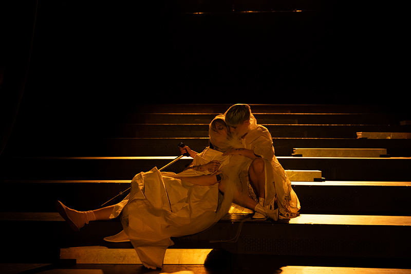 Two performers dressed in flowing light-colored costumes sit closely together on dimly lit steps, illuminated by warm golden stage lighting.