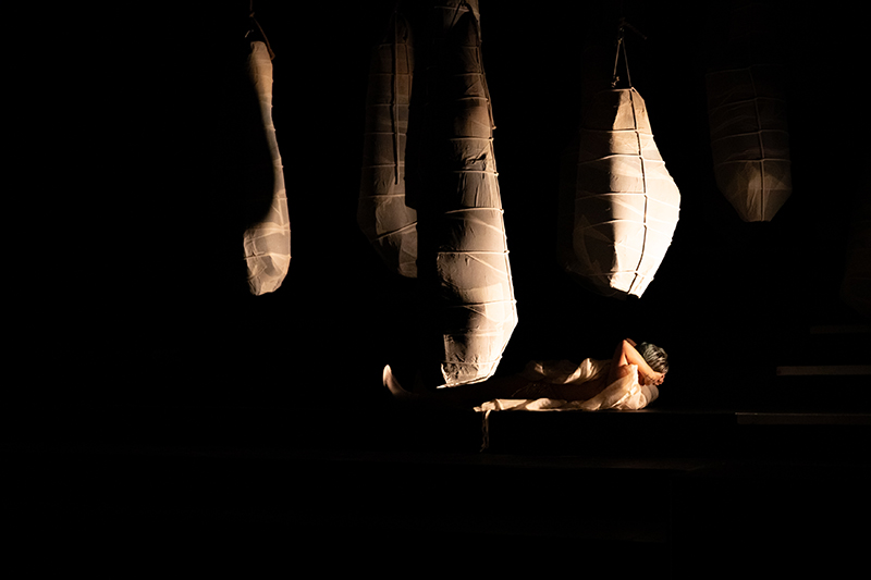 A performer lies on a dimly lit stage beneath several large, cocoon‑like hanging forms that glow softly in the darkness.