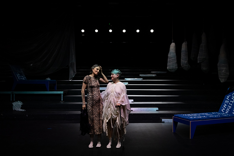 Two performers stand side by side on a dimly lit stage, dressed in contrasting textured costumes amid a backdrop of steps, hanging forms, and blue stage furniture.