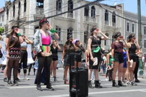 A group of performers stand in rows outside on the street. They are blindfolded and bring a hand to their mouths. They are yelling.