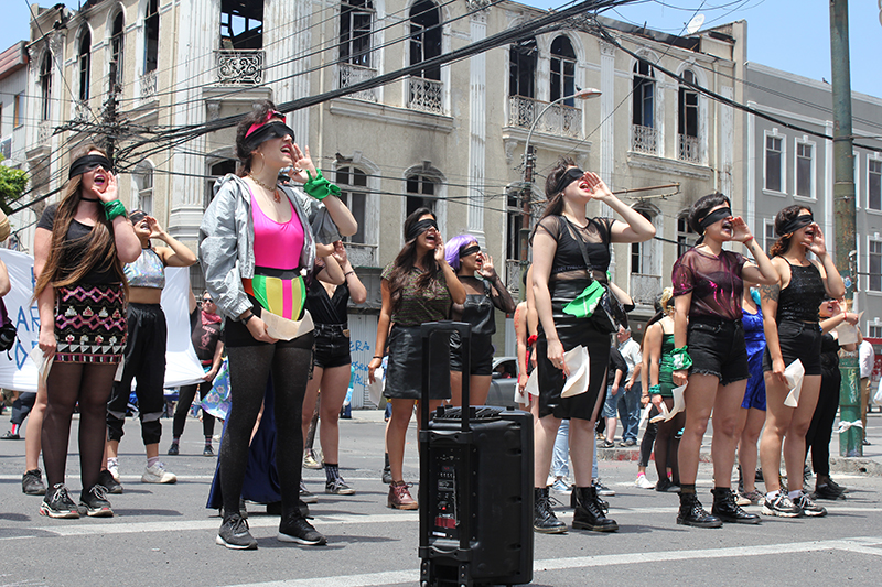 A group of performers stand in rows outside on the street. They are blindfolded and bring a hand to their mouths. They are yelling.