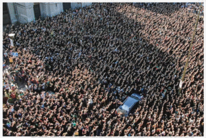 A massive crowd as seen from above surrounds a car. Everyone in the crowd has their arms lifted.