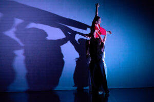 A group of dancers performs a dramatic contemporary lift with one dancer walking on a wall supported by the others, casting expressive shadows that amplify the visual impact.
