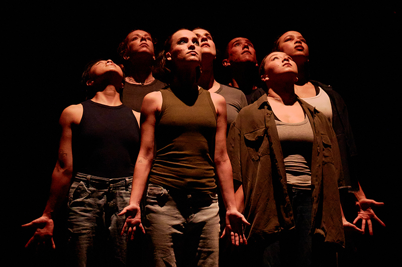 A group of performers stands in a dramatic stage formation with expressive, open-handed poses under focused lighting.