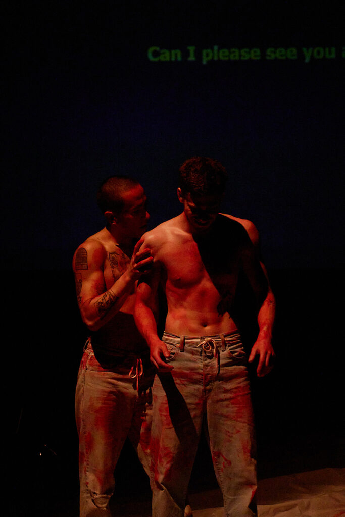 Two shirtless performers onstage under red light, one stands behind the other and touches their shoulder.