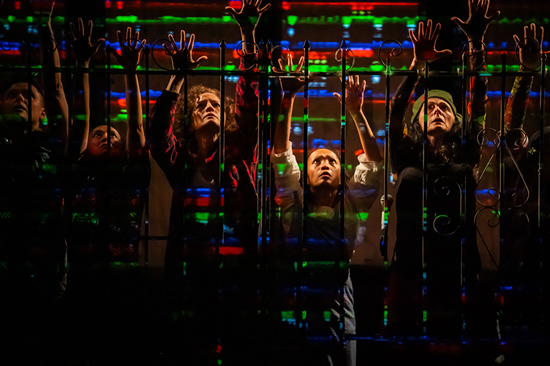 A group of people stand behind a metal fence with their hands raised, illuminated by colorful horizontal light patterns in red, green, and blue.
