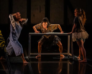 Three performers are engaged in a dramatic scene on stage, with one leaning against a rectangular metal table in a wide stance while the others pose expressively on either side, all barefoot and wearing eclectic costumes.