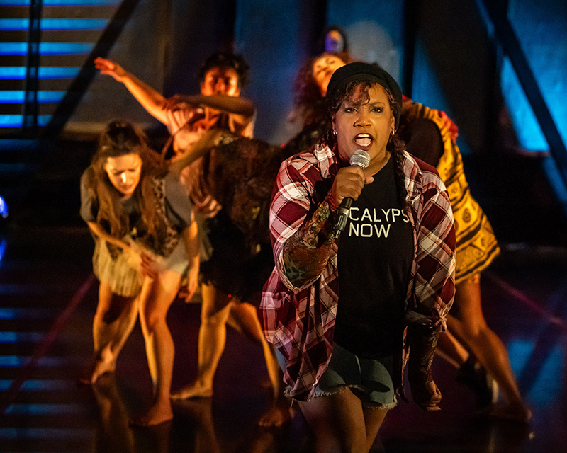 A person wearing a plaid shirt and a black T-shirt that reads “APOCALYPSE NOW” holds a microphone in the foreground, while a group of barefoot performers in patterned costumes pose dynamically behind them on a dimly lit stage.