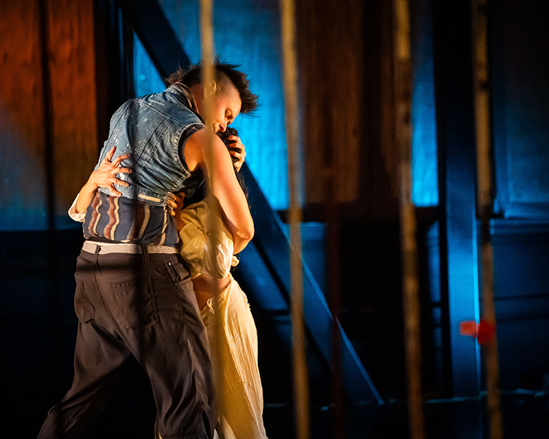One person is lifting another in an embrace on a dimly lit stage with a wooden backdrop and blue lighting accents.