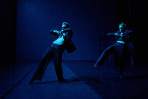 Two dancers move in dramatic, angular poses under deep blue stage lighting, their jackets pulled open as they lean and balance in dynamic motion.