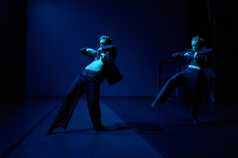 Two dancers move in dramatic, angular poses under deep blue stage lighting, their jackets pulled open as they lean and balance in dynamic motion.