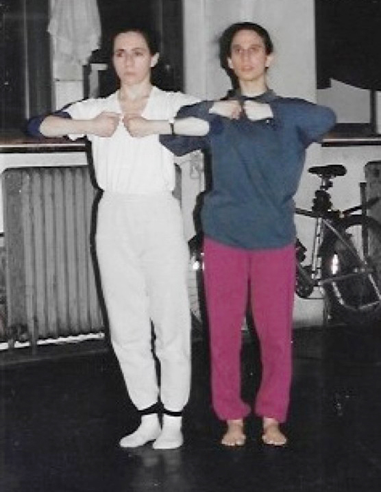 Two dancers stand in a studio with arms linked across their chests.