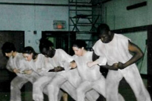 Five dancers in a high school gym link arms and lunge. They are wearing white.