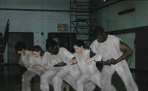 Five dancers in a high school gym link arms and lunge. They are wearing white.