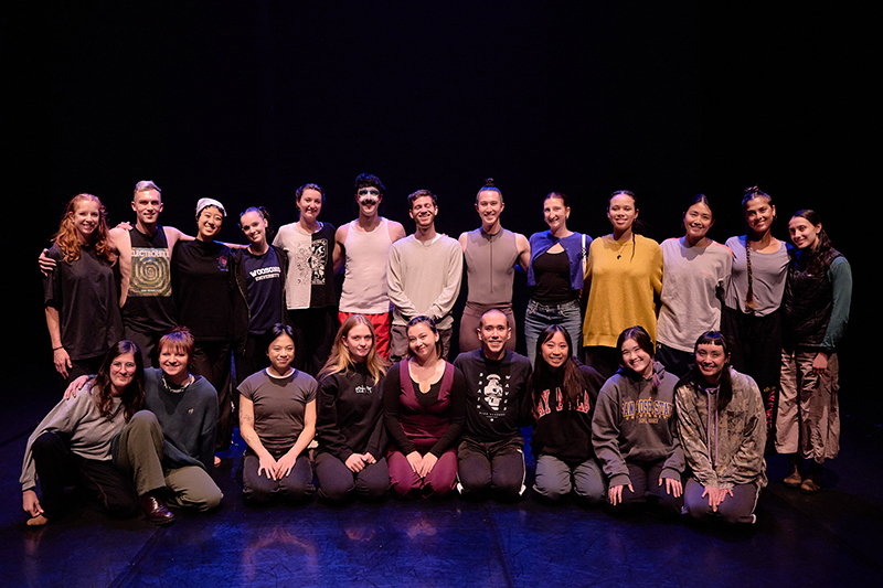 A large group of performers poses together on a dimly lit stage, arranged in two rows for a cast photo.