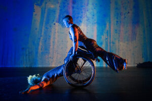 A moment from Kinetic Light’s The Next TiMes. Laurel Lawson holds their body in a diagonal line as they balance atop Alice Sheppard’s wheels and shins. Alice supports from below as they lay back, arms and torso on the ground, hips lifted. Alice is a multiracial Black woman with short curly hair, Laurel is a white person with very short cropped hair; they both wear shimmery sleeveless bodysuits. Behind them, a projection of soft blues, purples, and greens looks like rain on a windowpane. Photo by Cherylynn Tsushima.