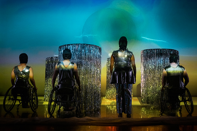 The Next TiMes. Four performers dressed in silver, with black caps, pause with their backs to the audience, three people in chairs and one standing. They gaze at four shimmering towers of silver fringe. Photo by Cherylynn Tsushima.