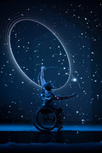 A moment from Kinetic Light’s The Next TiMes. Laurel Lawson glides across the stage, curved arms lifted and mirroring not only the large silver hoop that encircles their shoulders but also the huge ring that appears in a starry sky behind them. Laurel is a white person with cropped hair. Photo by Cherylynn Tsushima.