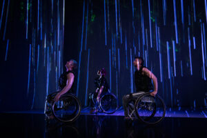 The Next TiMes. Laurel Lawson, Alice Sheppard, and Tatiana Cholewa wheel across the stage, gazing up. They all wear silver pants and shimmery black tops. In the background, a projection of long blue lines and dappled green reflections of light. Photo by Cherylynn Tsushima.