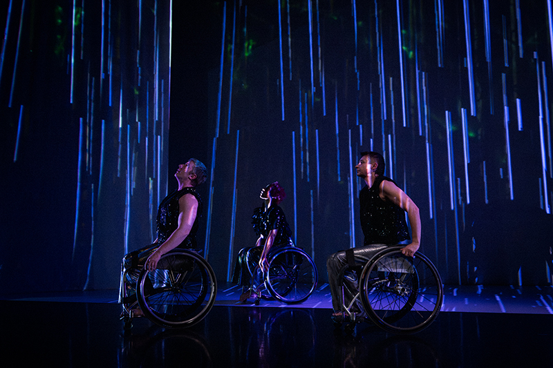 The Next TiMes. Laurel Lawson, Alice Sheppard, and Tatiana Cholewa wheel across the stage, gazing up. They all wear silver pants and shimmery black tops. In the background, a projection of long blue lines and dappled green reflections of light. Photo by Cherylynn Tsushima.
