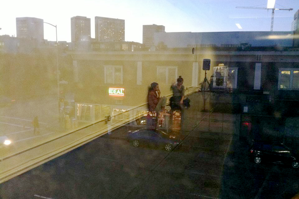 A reflection in a window shows two people sitting in a studio near a speaker setup as the cityscape glows at dusk.