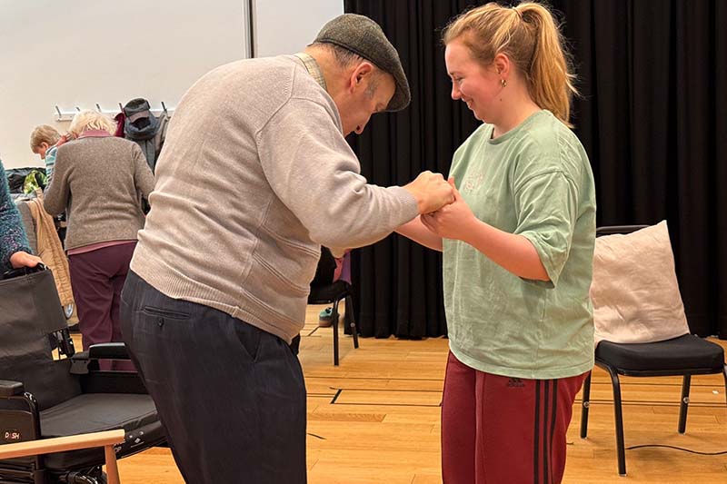 A young woman supports an older man in a gentle partnered movement exercise inside a studio space.