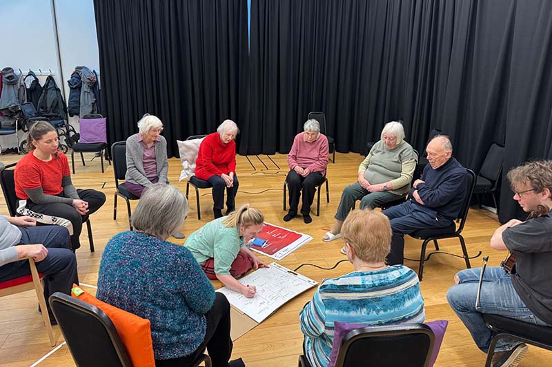 A group of people sit in a circle in a studio while one person kneels in the center drawing on a large sheet of paper.