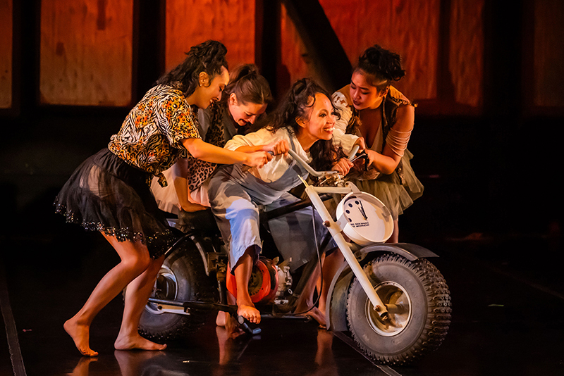 Four performers crowd together on a small motorcycle onstage.