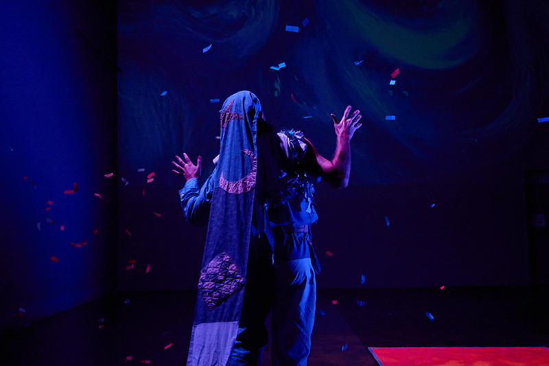 A performer wearing a veil and facing upstage with hands extended under dramatic blue lighting as colorful confetti falls around them.