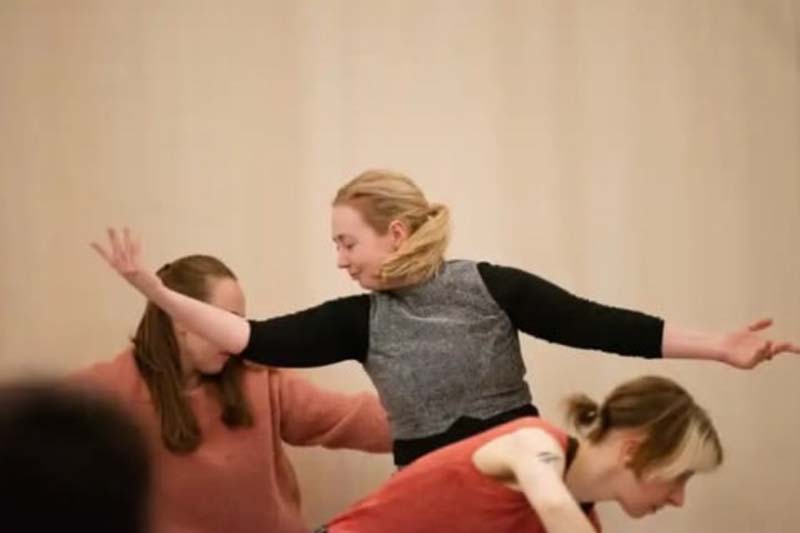 Two people surround a third person as they extend their arms wide in a balanced, expressive movement.