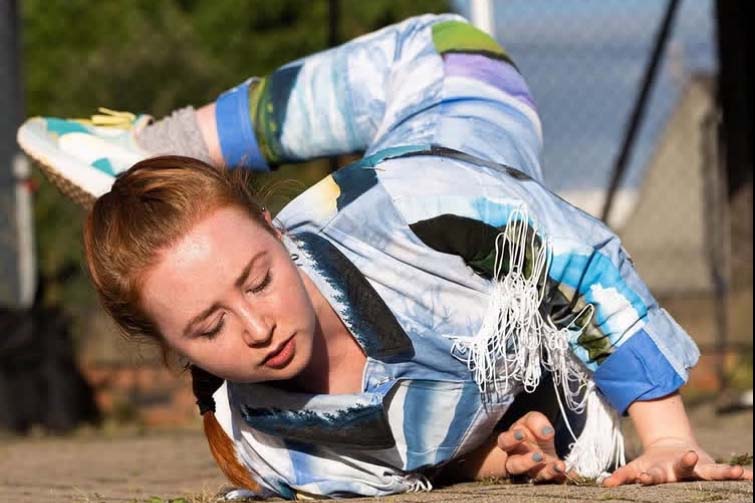 A person in a colorful outfit performs a low, grounded movement on an outdoor surface with one leg lifted behind them.
