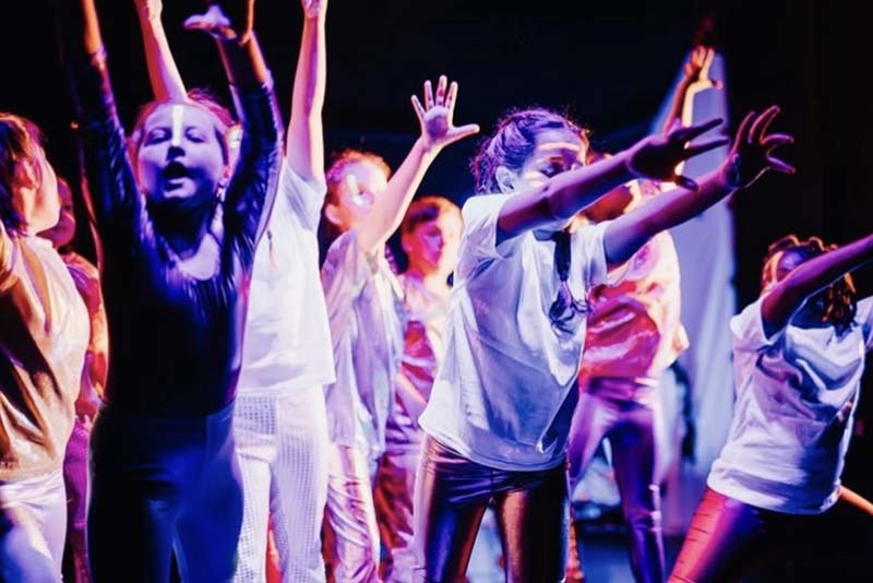 A group of children raises their arms in expressive, energetic movement under vibrant stage lighting.