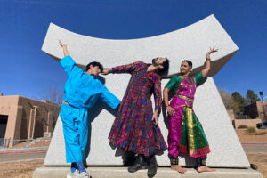 Three people in vibrant, flowing outfits strike expressive poses together in front of a large modern sculpture under a bright blue sky.