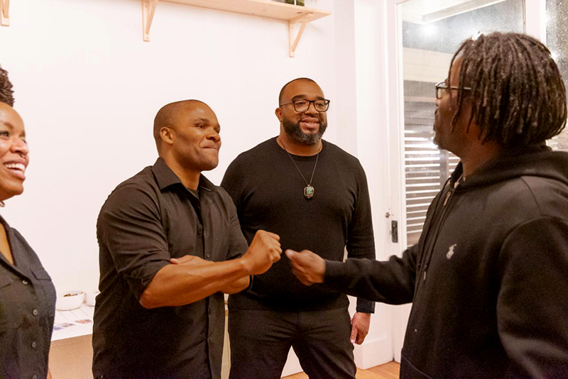 Four people stand together indoors as two of them greet each other with a friendly fist bump.
