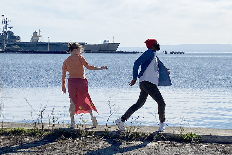 Two people dance playfully along a waterfront path with a large ship in the background.