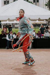 A dancer in vibrant, patterned regalia performs an energetic hoop dance in an outdoor event space.