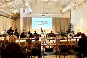 An audience sits in a warm, modern event space as a speaker presents onstage before a large screen displaying “International Museum of Dance.”