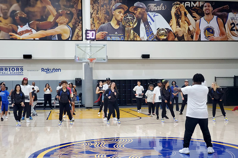 A group of people participates in a lively dance class on a professional basketball court, led by an instructor facing them.