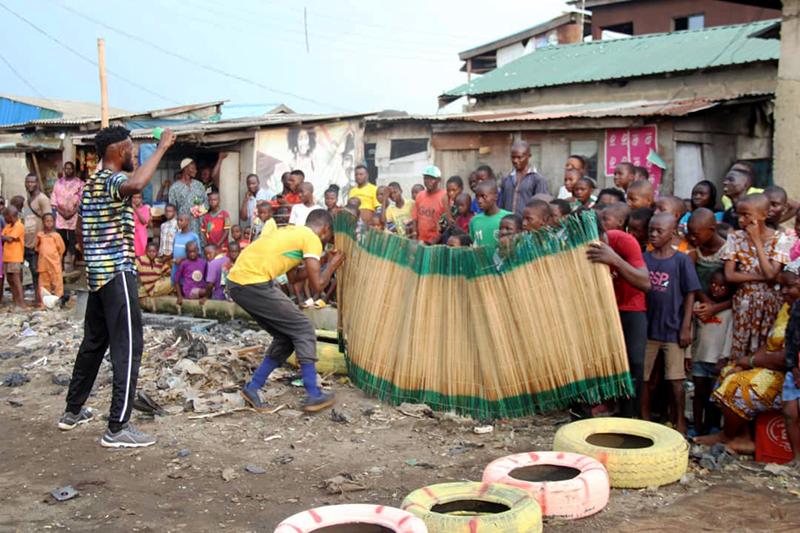 Performers engage in a lively outdoor routine while a crowd of children and adults gathered in a neighborhood setting watches with interest behind a tall woven barrier.