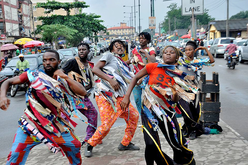 A group of dancers wearing colorful, ribbon-adorned costumes performs energetic movements along a busy urban roadside.