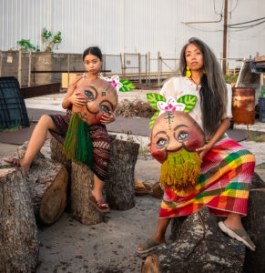 A Filipino brown mother (Ani Gavino) sits next to her teen daughter (Malaya Ulan); each one holds a Papier-mâché puppet mask created by artists, Kitchen Table Puppet Press. The masks symbolize ""sweet potato/kamote ancestors"" native to the Philippines. This photo is taken outside the Arts & Culture Lab in Houston, TX, one of AniMalayaworks' partner organizations.