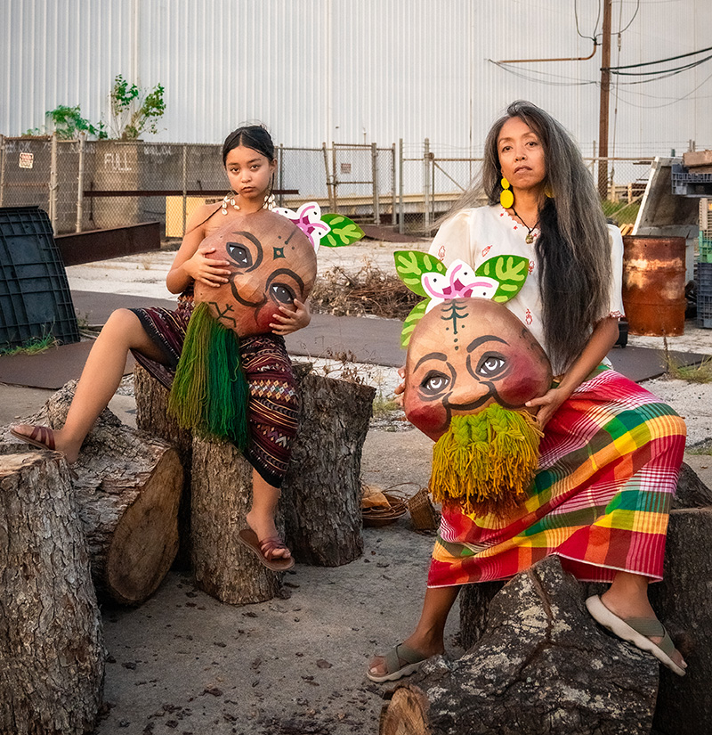 A Filipino brown mother (Ani Gavino) sits next to her teen daughter (Malaya Ulan); each one holds a Papier-mâché puppet mask created by artists, Kitchen Table Puppet Press. The masks symbolize ""sweet potato/kamote ancestors"" native to the Philippines. This photo is taken outside the Arts & Culture Lab in Houston, TX, one of AniMalayaworks' partner organizations. 