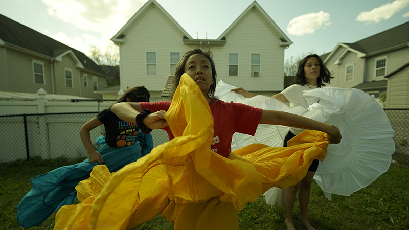 A Filipino brown woman (Ani Gavino) whips her yellow skirt in a dance form called Bomba (an Afro-Puertorican dance) along with two other dancers (Athena Turek and Malaya Ulan). They are rehearsing in a backyard. 