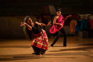 Ani and Malawa perform expressive, angular movements on a wooden stage, wearing patterned red garments over dark clothing under warm theatrical lighting with fabric props in the background.