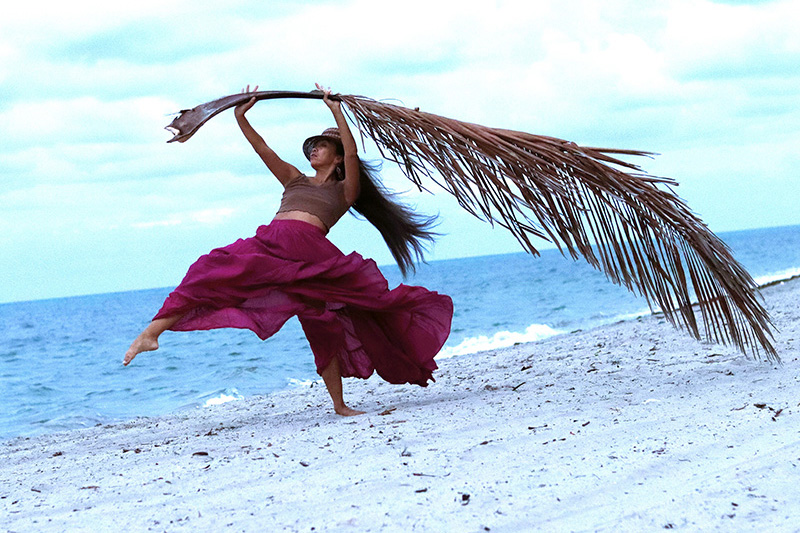 Ani leaps along a sandy shoreline, wearing flowing magenta clothing and lifting a long palm frond overhead as the ocean and pale blue sky stretch behind.