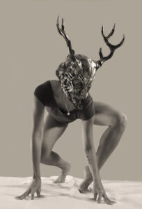 Ani crouches and balances on a light fabric surface, wearing a sculptural antlered mask and a leotard, posed against a plain backdrop with strong, dramatic contrast.