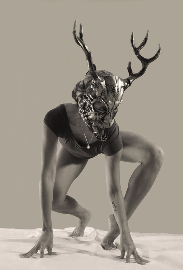 A woman crouches in a sepia tone photo. She wears a metallic deer head. 